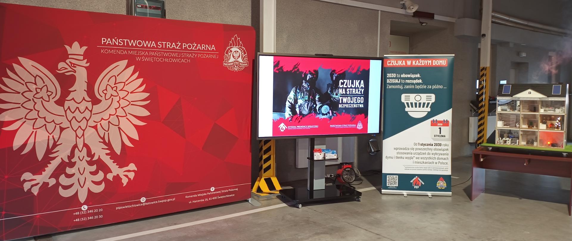 Zdjęcie przedstawia profesjonalne zaplecze edukacyjne przygotowane przez Komendę Miejską Państwowej Straży Pożarnej w Świętochłowicach, dedykowane kampanii społecznej „Czadowa Straż”. Na pierwszym planie widoczna jest duża czerwona ścianka z godłem Polski, a obok niej ustawiono ekran multimedialny oraz baner informujący o obowiązku montażu czujek dymu i tlenku węgla od 2030 roku. Uzupełnieniem stanowiska jest makieta domku jednorodzinnego, z której wydobywa się dym, co służy do praktycznego zademonstrowania zagrożeń pożarowych oraz skuteczności urządzeń alarmowych w warunkach domowych.