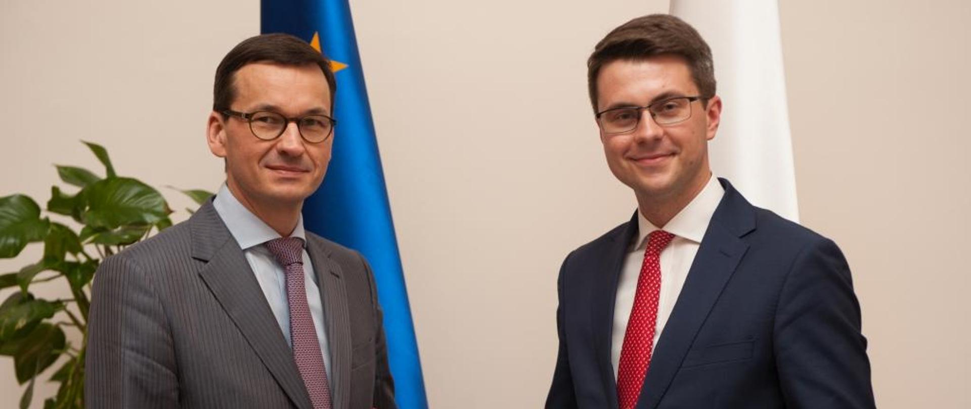 Premier Morawiecki wręcza dokument Piotrowi Mullerowi, w tle flagi Polski i UE