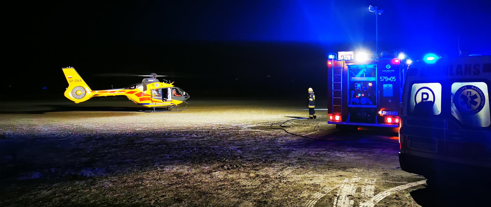 Zabezpieczenie lądowiska dla LPR przez strażaków. Na zdjęciu z lewej strony widać śmigłowiec Lotniczego Pogotowia Ratunkowego, po prawej widoczny wóz strażacki z włączonym oświetleniem oraz karetka. 
