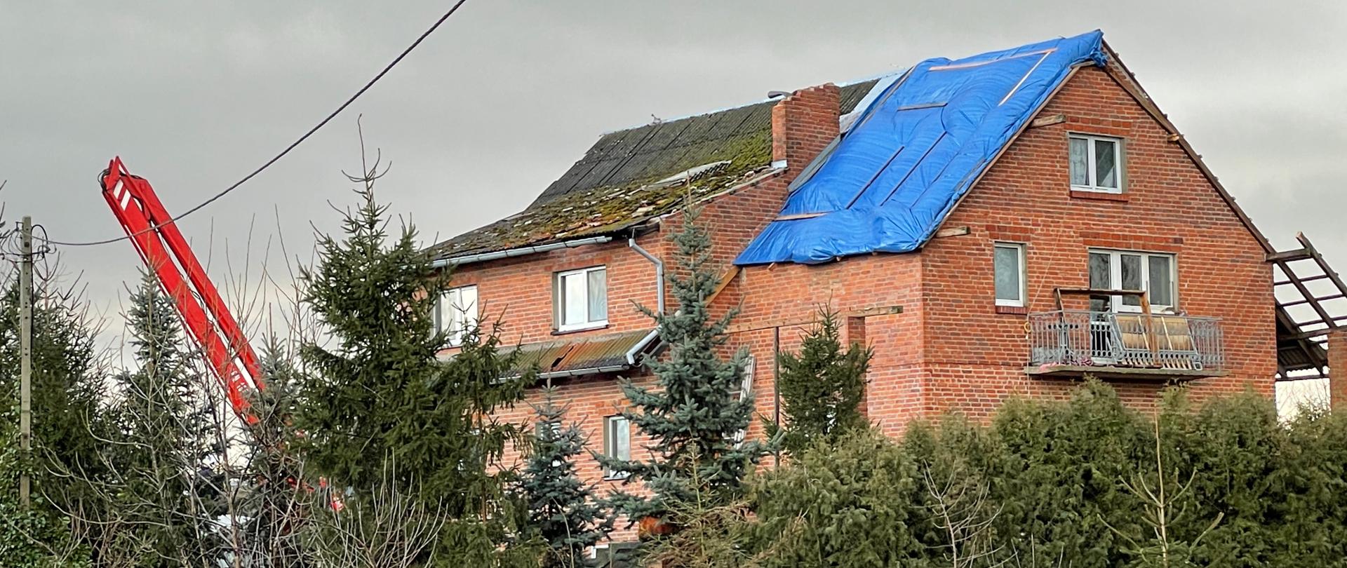 Budynek mieszkalny z czerwonej cegły na 1/3 dachu budynku założona niebieska plandeka przed budynkiem drzewa iglaste zboku widoczne ramie podnośnika hydraulicznego