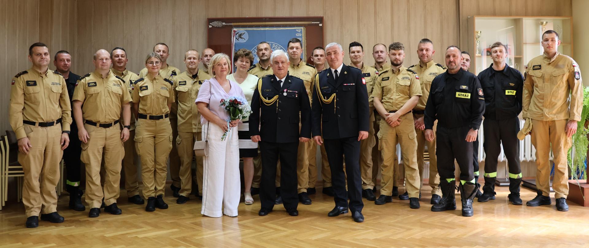 Na zdjęciu widoczna jest duża grupa osób pozujących wspólnie do pamiątkowej fotografii w sali o drewnianej podłodze podczas uroczystości przejścia na zaopatrzenie emerytalne. Większość stanowią strażacy w mundurach służbowych koloru beżowego, kilku ubranych jest w czarne lub jasne mundury koszarowe z naszywką „STRAŻ”. Na środku stoją dwie kobiety w cywilnych ubraniach – jedna z nich trzyma bukiet kwiatów – oraz Komendant i emeryt w czarnych galowych mundurach strażackich z odznaczeniami i złotymi sznurami naramiennymi. W tle widać gablotę z trofeami oraz sztandar, co podkreśla uroczysty charakter spotkania.