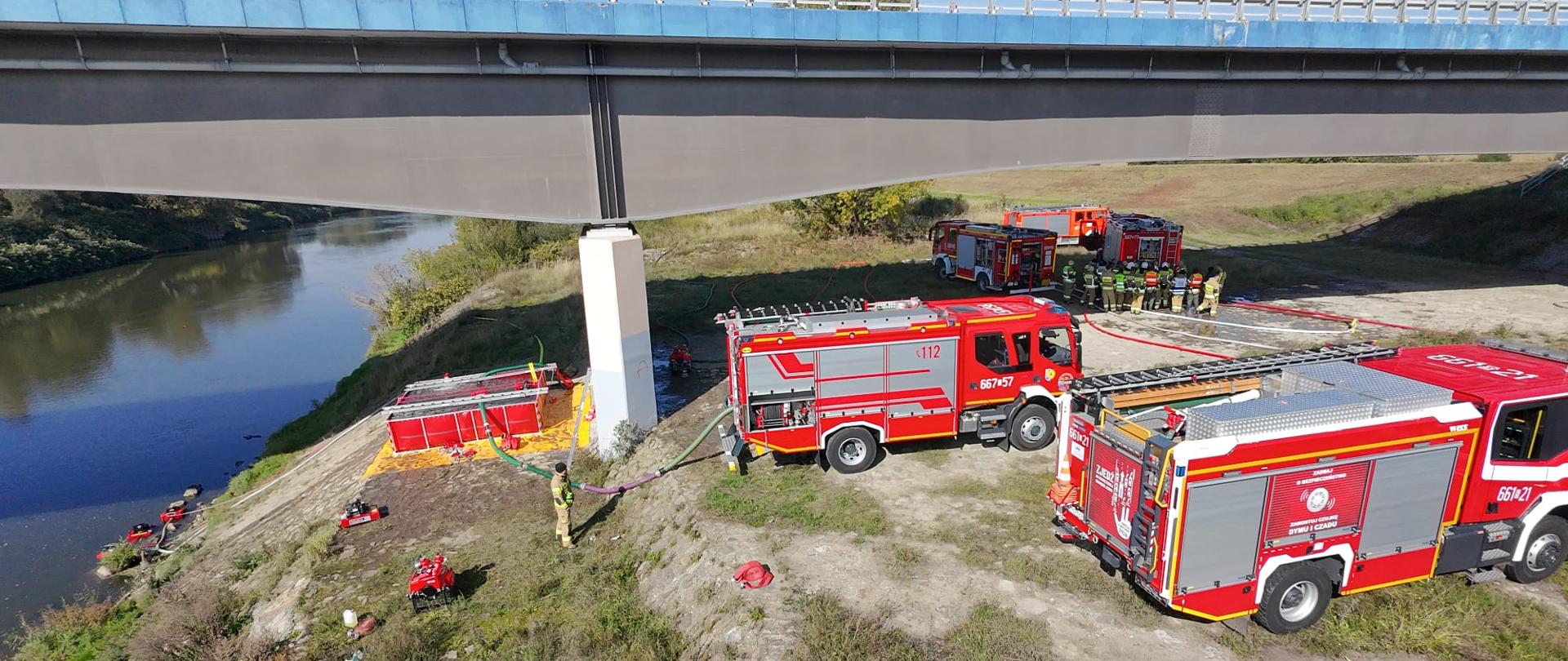 Samochody pożarnicze stoją nad ciekiem wodnym pobierając z niego wodę, w tle strażacy i pojazdy pożarnicze.