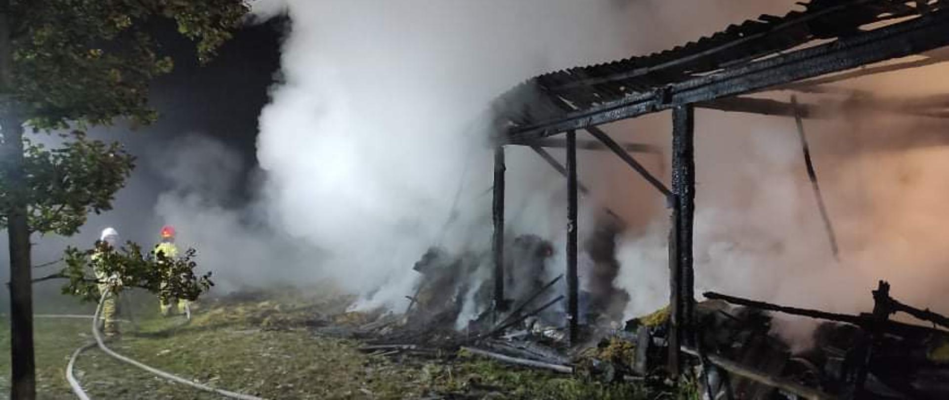 Na zdjęciu widać spaloną konstrukcję stodoły. Wewnątrz pali się słoma i siano. Nad budyniem z budynku wydobywa się zadymienie. Po lewej stronie zdjęcia znajduje się dwóch strażaków, którzy gaszą pożar wodą. Teren akcji w nocy oświetla specjalna najaśnica.