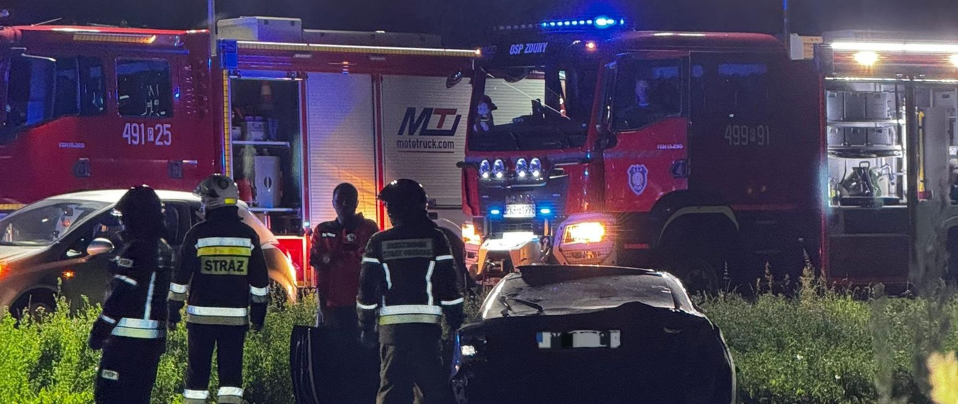 Zdjęcie przedstawia strażaków podczas wypadku drogowego w porze nocnej, w tle wozy strażackiej.