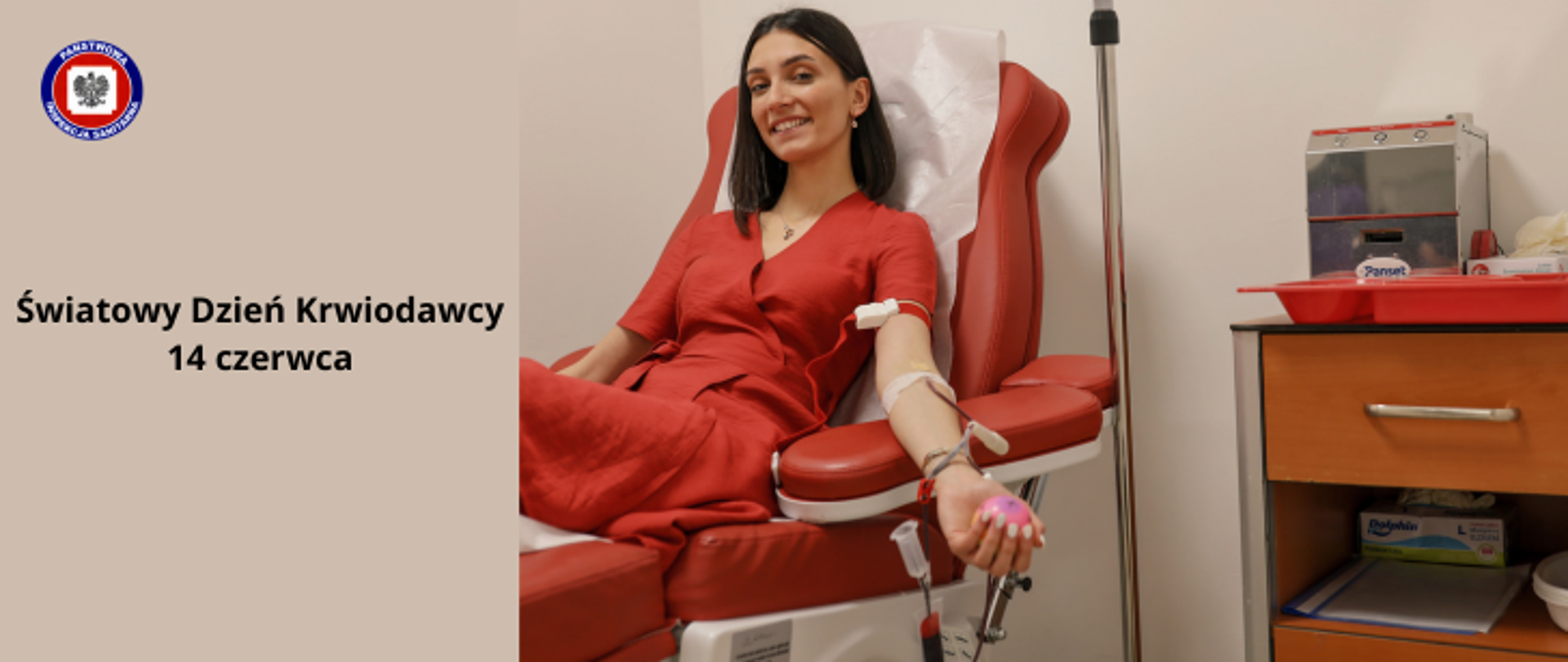 Po prawej uśmiechnięta brunetka w czerwonej sukience siedząca na czerwonym fotelu w gabinecie lekarskim podczas czynności z transfuzją krwi. Po lewej na jasnobrązowym tle ciemny napis Światowy Dzień Krwiodawcy. W lewym górnym rogu logo Państwowej Inspekcji Sanitarnej.