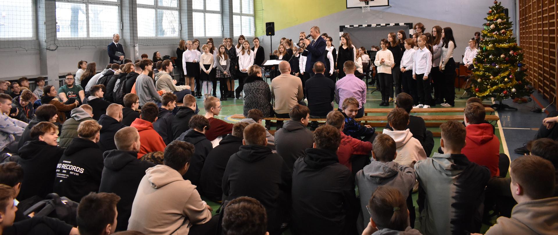 na zdjęciu w sali gimnastycznej na pierwszym planie widnieje społeczność szkolna ZST w Jaśle, która witana jest przez dyrektora ZST. W tle zdjęcia widoczny jest chór szkolny. Kolorystyka zdjęcia jest biało-żółto-czerwono-brązowo-czarna.