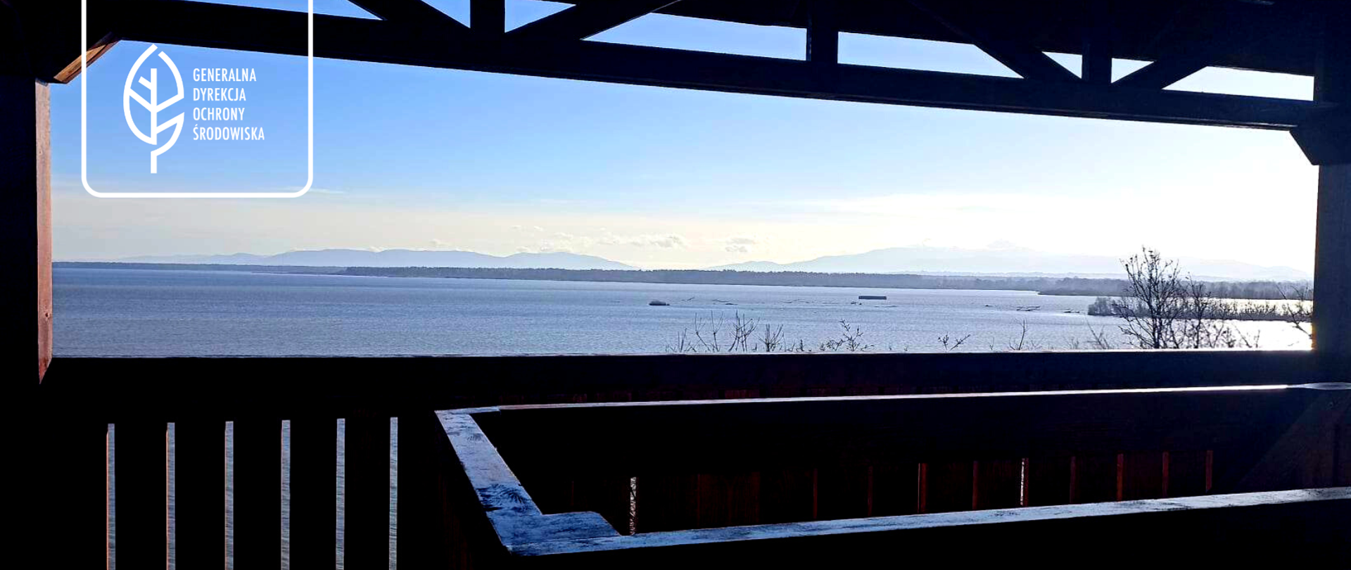Widok z drewnianej wieży widokowej. Krajobraz jeziora, pagórków o krzaków. Niebo zachmurzone. W lewym górnym roku logo Generalnej Dyrekcji Ochrony Środowiska (biały liść).