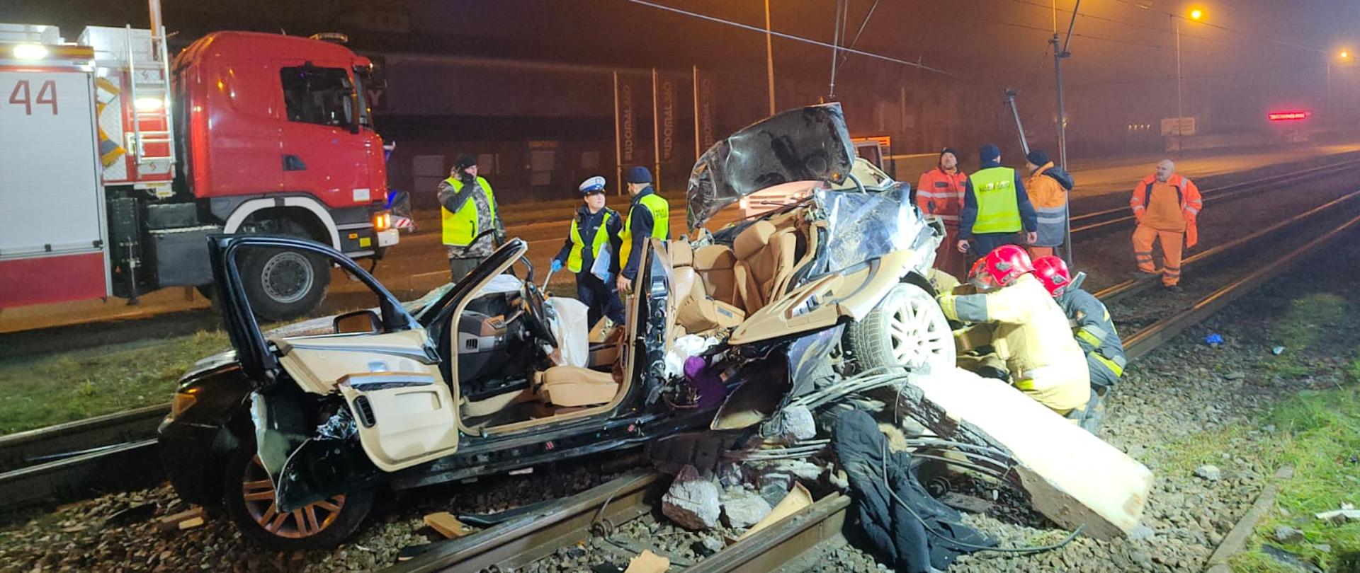 Wrak samochodu stojącego na torach tramwajowych, w tle samochód pożarniczy oraz funkcjonariusze PSP i Policji