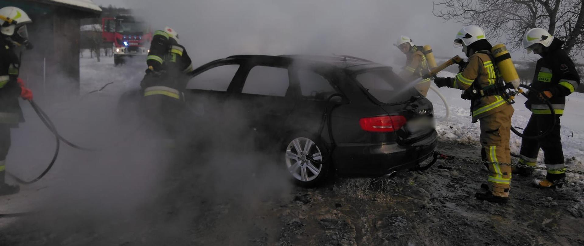 Pięciu strażaków gasi pożar samochodu obok budynku. Z samochodu unosi się dym. Na ziemi widać warstwę białego oraz roztopionego śniegu.