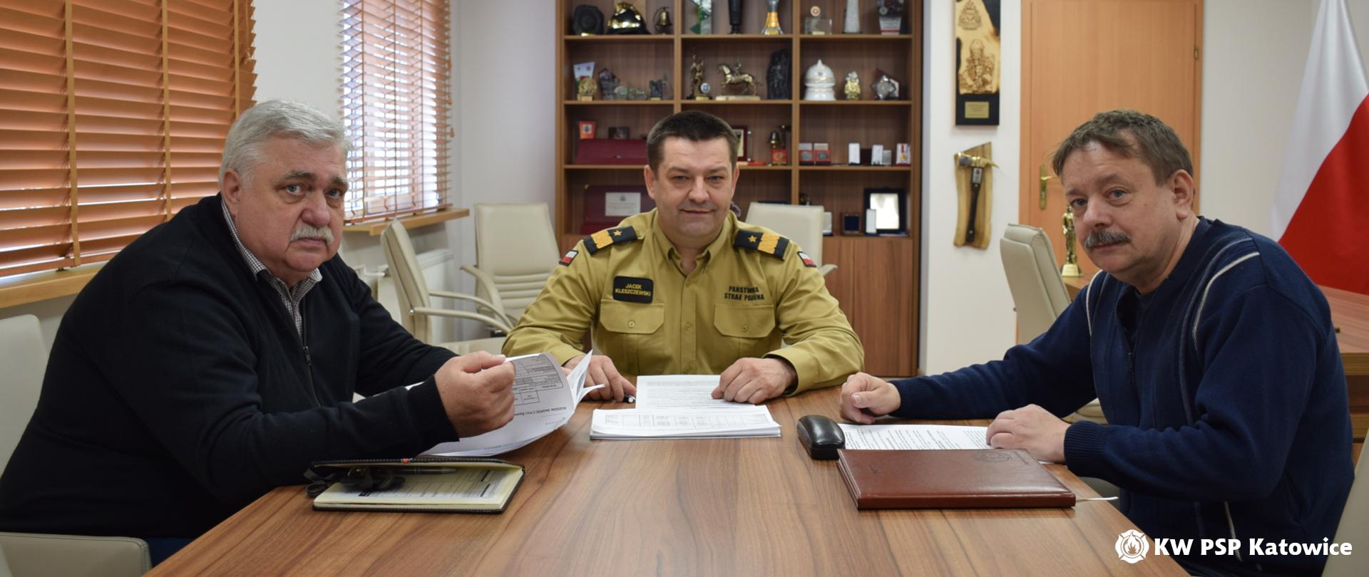 W gabinecie komendanta wojewódzkiego siedzą przy stole: na wprost śląski komendant wojewódzki PSP nadbryg. Jacek Kleszczewski, po prawej stronie przewodniczący Międzyregionalnej Śląskiej Sekcji Pożarnictwa NSZZ „Solidarność” Panem Damianem Kochoniem , po lewej przewodniczący Śląskiego Zarządu Wojewódzkiego Związku Zawodowego Strażaków „Florian” Panem Jarosławem Nowakiem. Na stole leżą dokumenty. Z tyłu widoczna gablota z pucharami i pamiątkami. 