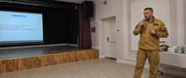 zdjęcie przedstawia strażaka w ubraniu służbowym z mikrofonem podczas referowania, w tle widoczny projektor