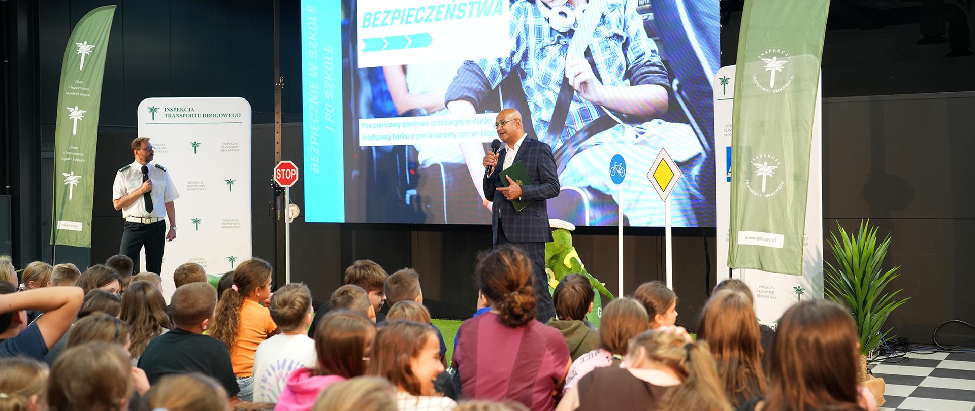 Zdjęcie od tyłu pod kątem, sala wypełniona siedzącymi na ławkach dziećmi, przed nimi mężczyzna w garniturze mówi do mikrofonu, za nim ścianka z napisem Pasy bezpieczeństwa.