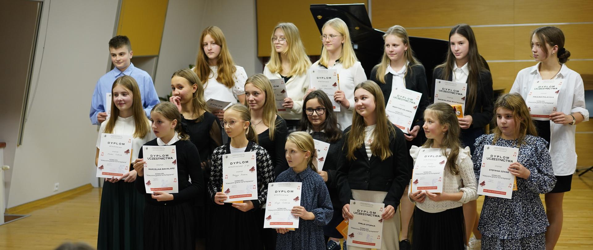 Na zdjęciu widać grupę nastoletnich uczestników konkursu stojących razem na scenie w sali muzycznej. To kilkanaście osób – głównie dziewczęta oraz jeden chłopiec – ustawionych w trzech rzędach. Wszyscy trzymają w rękach dyplomy udziału w konkursie. Młodzież jest elegancko ubrana: dziewczęta mają spódnice, sukienki lub eleganckie bluzki, a chłopiec jasną koszulę. Za nimi stoi czarny fortepian, a w tle widać jasne, gładkie ściany oraz żółte rolety w oknach. Uczestnicy wyglądają na uśmiechniętych i zadowolonych. Pozują do zdjęcia, stojąc blisko siebie, jak po zakończonym występie lub gali rozdania nagród. Cała scena ma uroczysty, szkolno-artystyczny charakter.