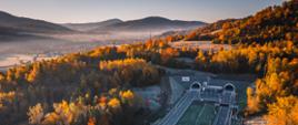 Widok z lotu ptaka na drogę S1 prowadzącą do wylotów tunelu, otoczoną jesiennym, górskim krajobrazem; w tle widać dolinę spowitą mgłą i pasma wzgórz oświetlone porannym słońcem