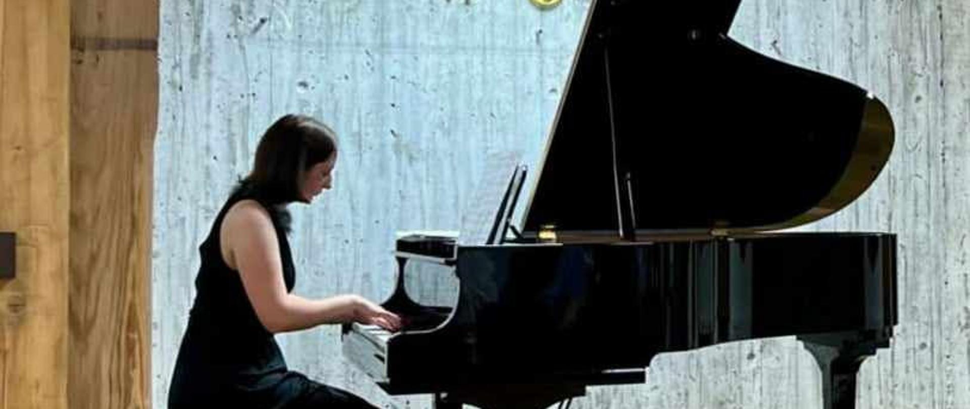 Zdjęcie kolorowe. Młoda pianistka w czarnej sukni gra na fortepianie. Drewniana scena. W tle napis Muzeum Pomorza Środkowego.