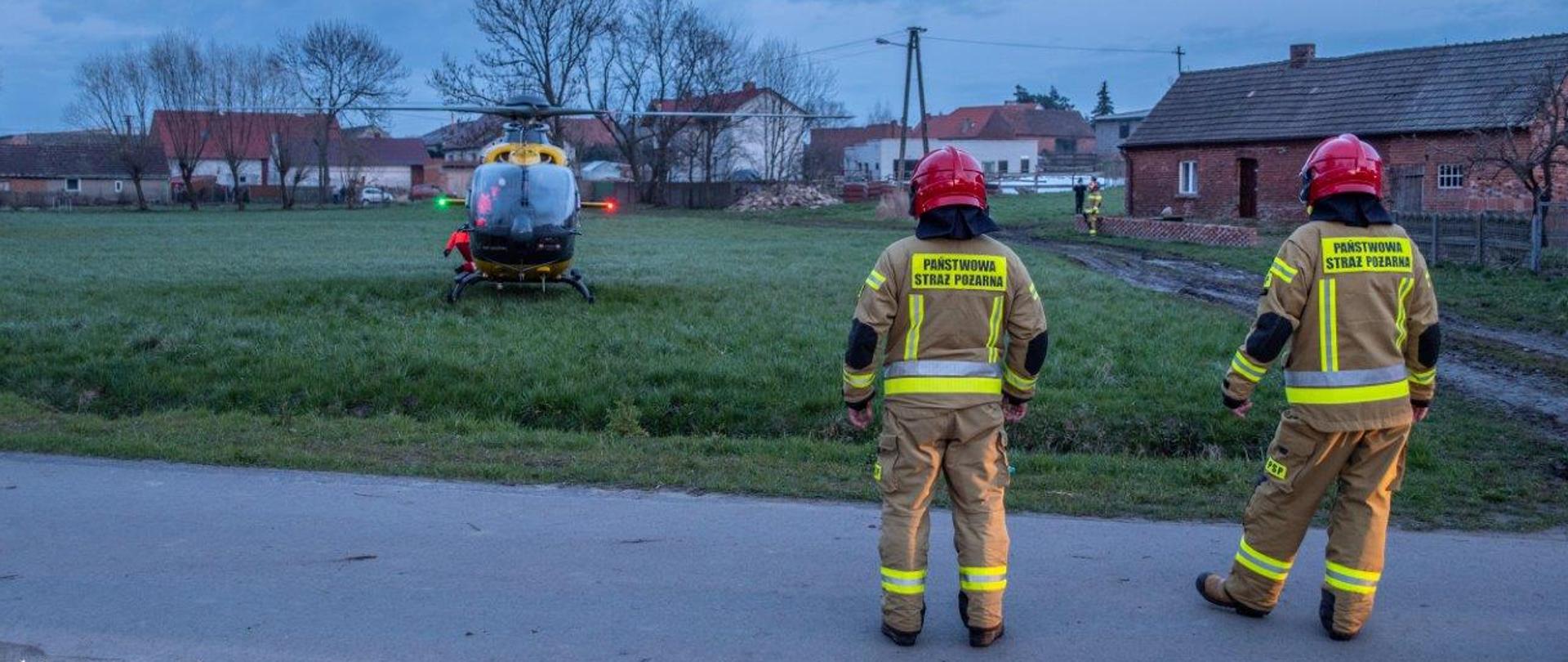 Na drodze przy polu uprawnym stoją strażacy, zabezpieczając start śmigłowca LPR. Śmigłowiec startuje do miejsca stacjonowania. 