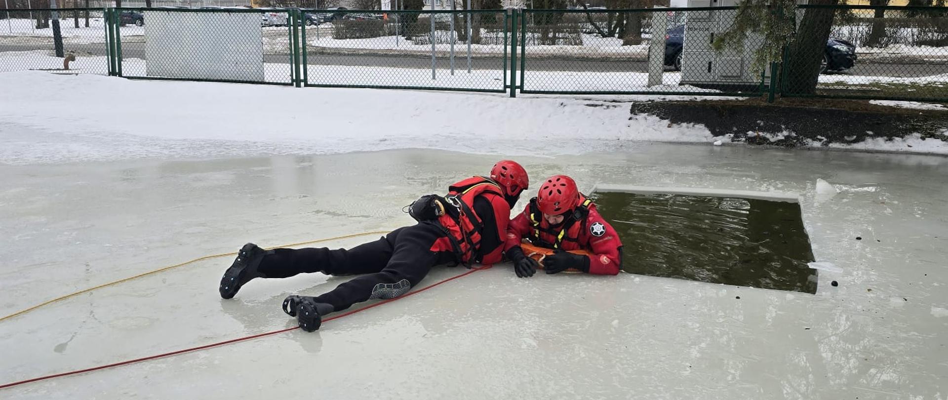 Ratownik w czerwonym kombinezonie i kasku czołga się po zamarzniętej tafli wody w stronę przerębla. Jest asekurowany linami. Obok niego, przy krawędzi lodu, znajduje się drugi ratownik.