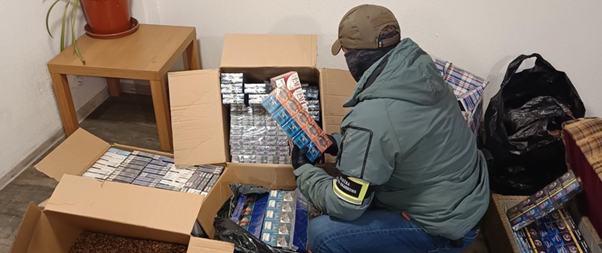 Funkcjonariusz trzyma w rękach karton z papierosami i ogląda papierosy w pudełkach. przed nim karton z tytoniem.