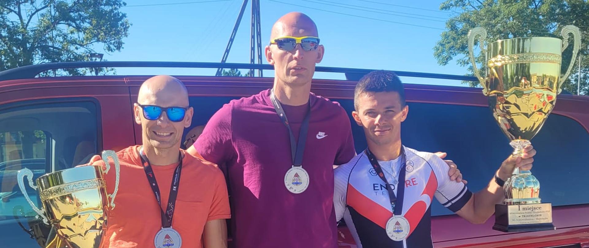 Zdjęcie wykonano w ciągu dnia, przedstawia trzech strażaków ubranych w ubrania sportowe z medalami na szyi i z pucharami w rękach. W tle stoi samochód strażacki typu bus.