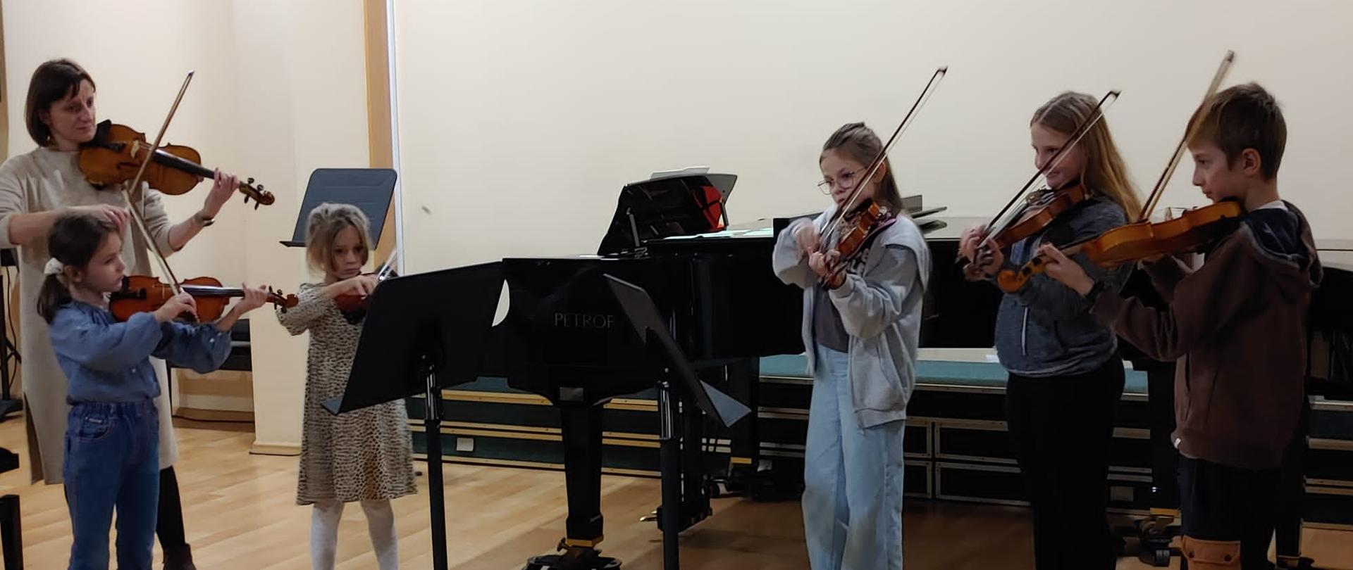 Po lewej stronie na zdjęciu widać panią która gra na skrzypcach a od lewej do prawej stoi pięciu uczniów przy pulpitach którzy też trzymają w rękach skrzypce i smyczek za nimi stoi fortepian a na ścianie logo szkoły PSM