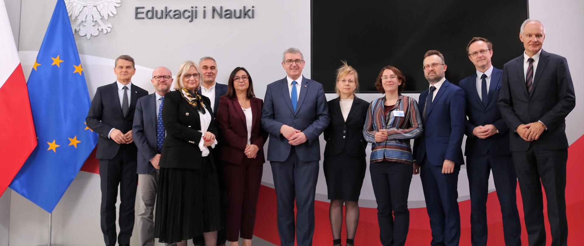 Zdjęcie zbiorowe - członkowie KPN stoją pod ścianą, za nimi flagi Polski i UE.