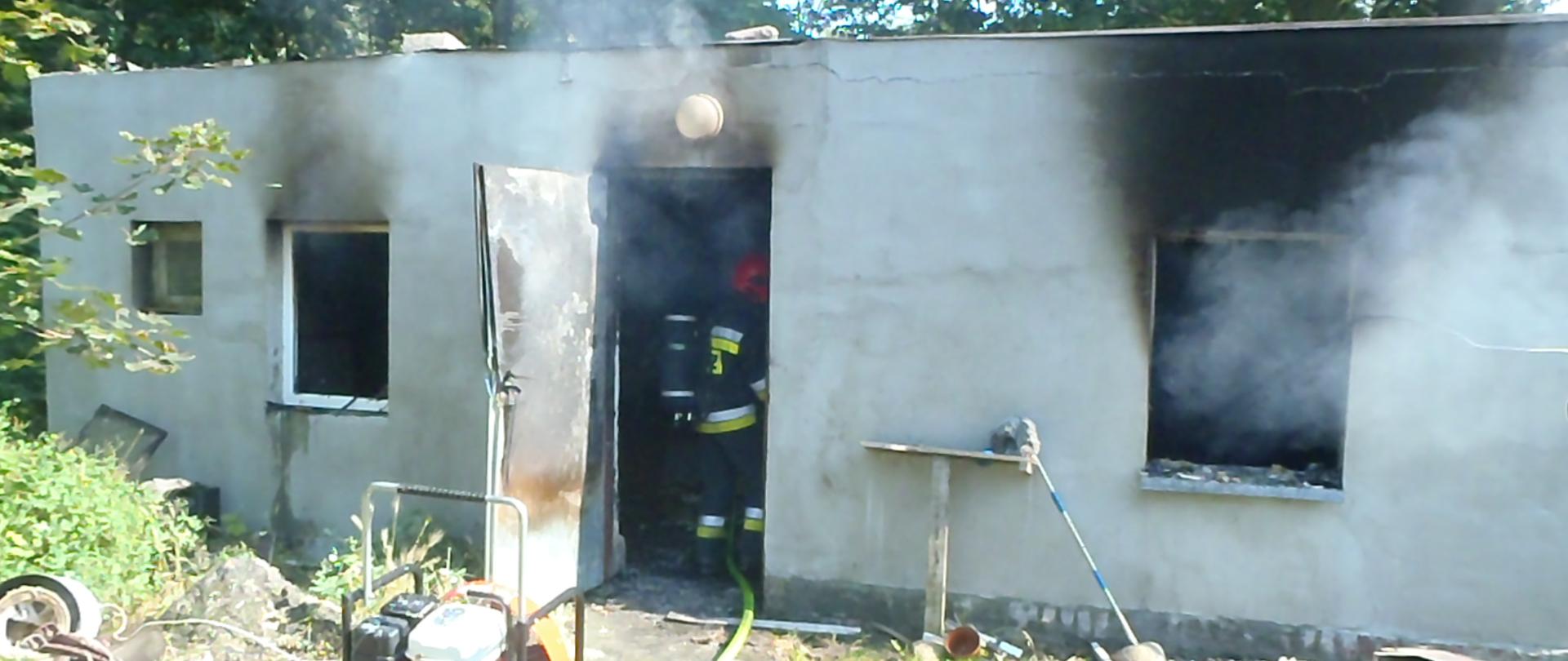 z parterowego budynku przed otwarte drzwi i wybite okno wydobywa się w tym w drzwiach w aparacie OUO stoi strażak prze budynkiem znajduje się agregat oddymiający 