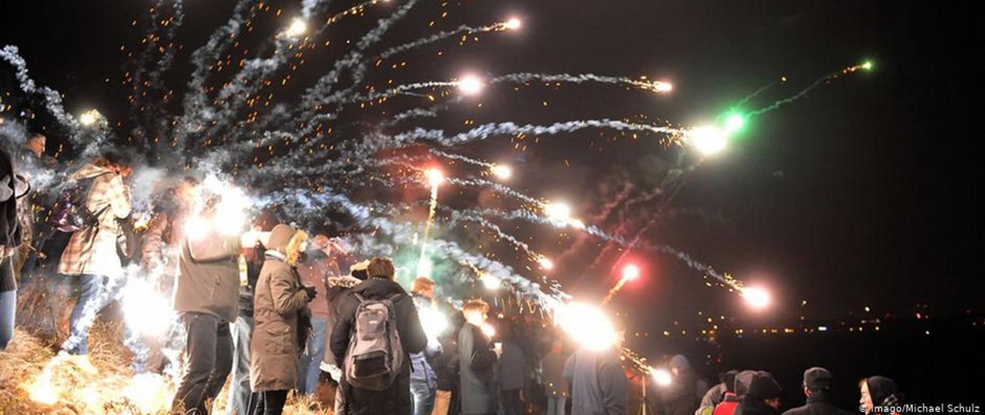 Strzelające w niebo fajerwerki oraz grupa ludzi stojąca i obserwująca rakiety.