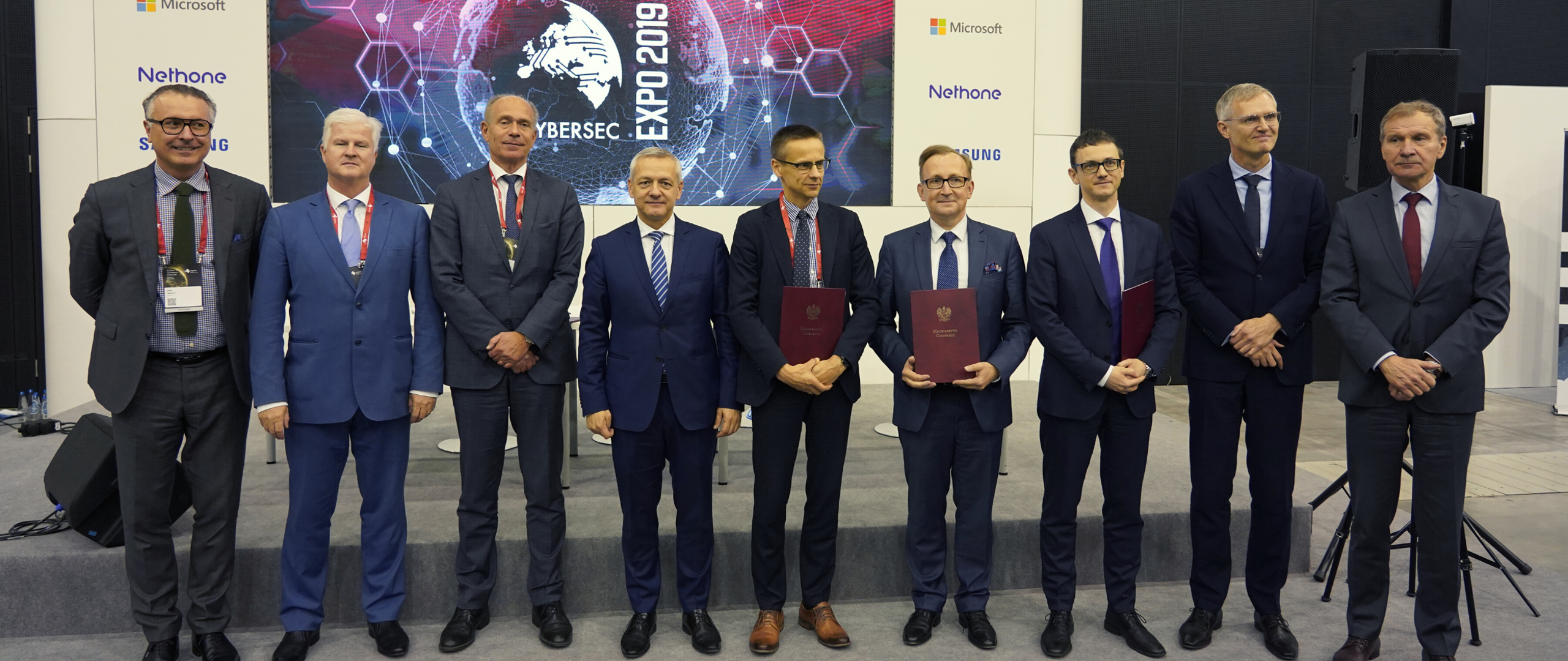 Dziewięciu mężczyzn pozuje do wspólnego zdjęcia na tle ścianki z logotypem wydarzenia