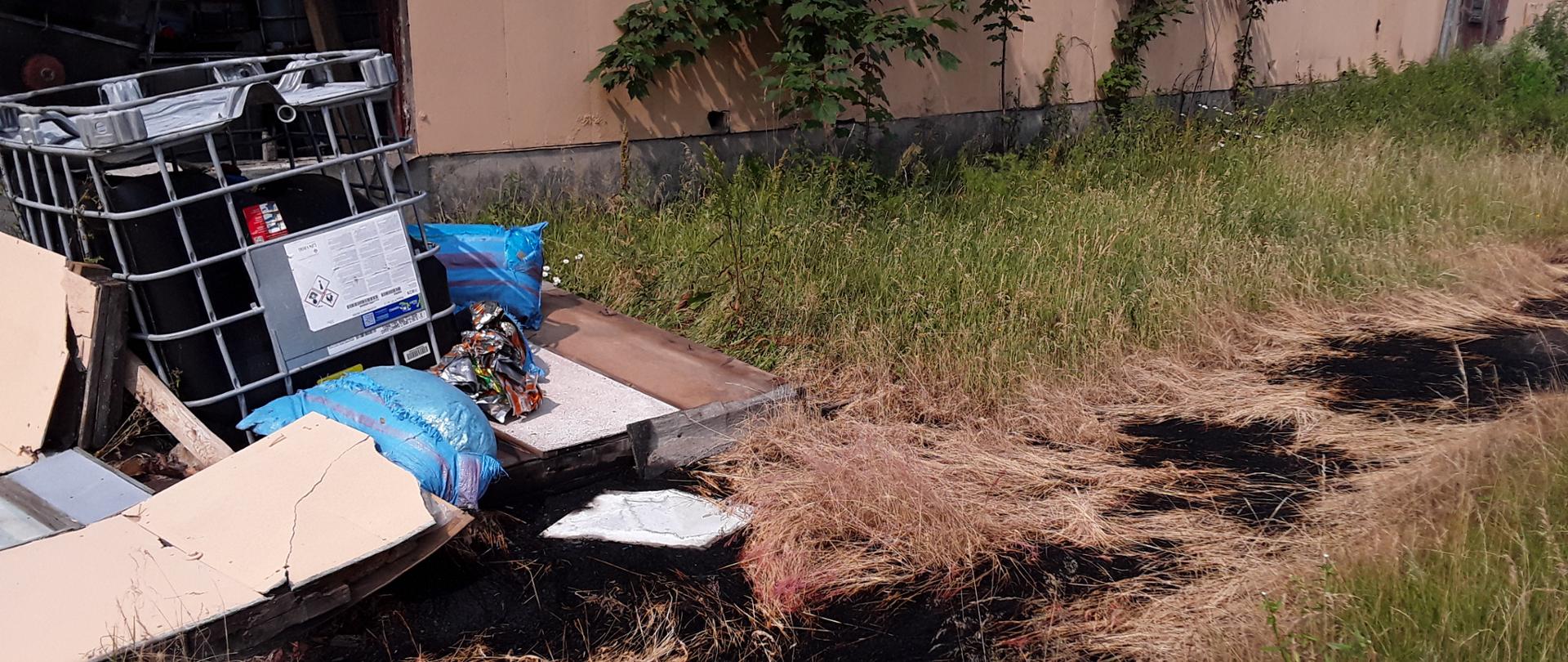 Przewrócony pojemnik o pojemności 1000 l na łąkę, na której jest pas wypalonej trawy o powierzchni kilku metrów kwadratowych. W tle ściana hali magazynowej.