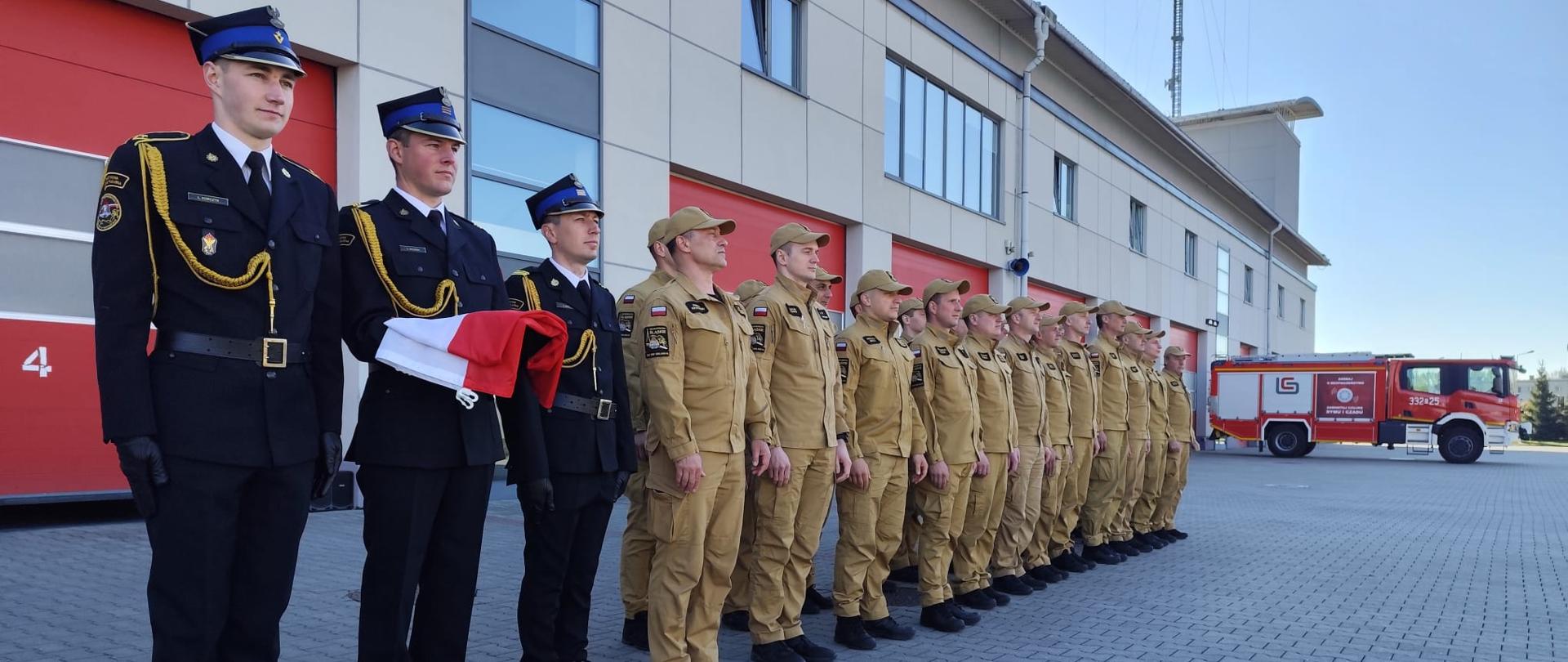 Strażacy w mundurach galowych i piaskowych stoją przed remizą obok wozu.