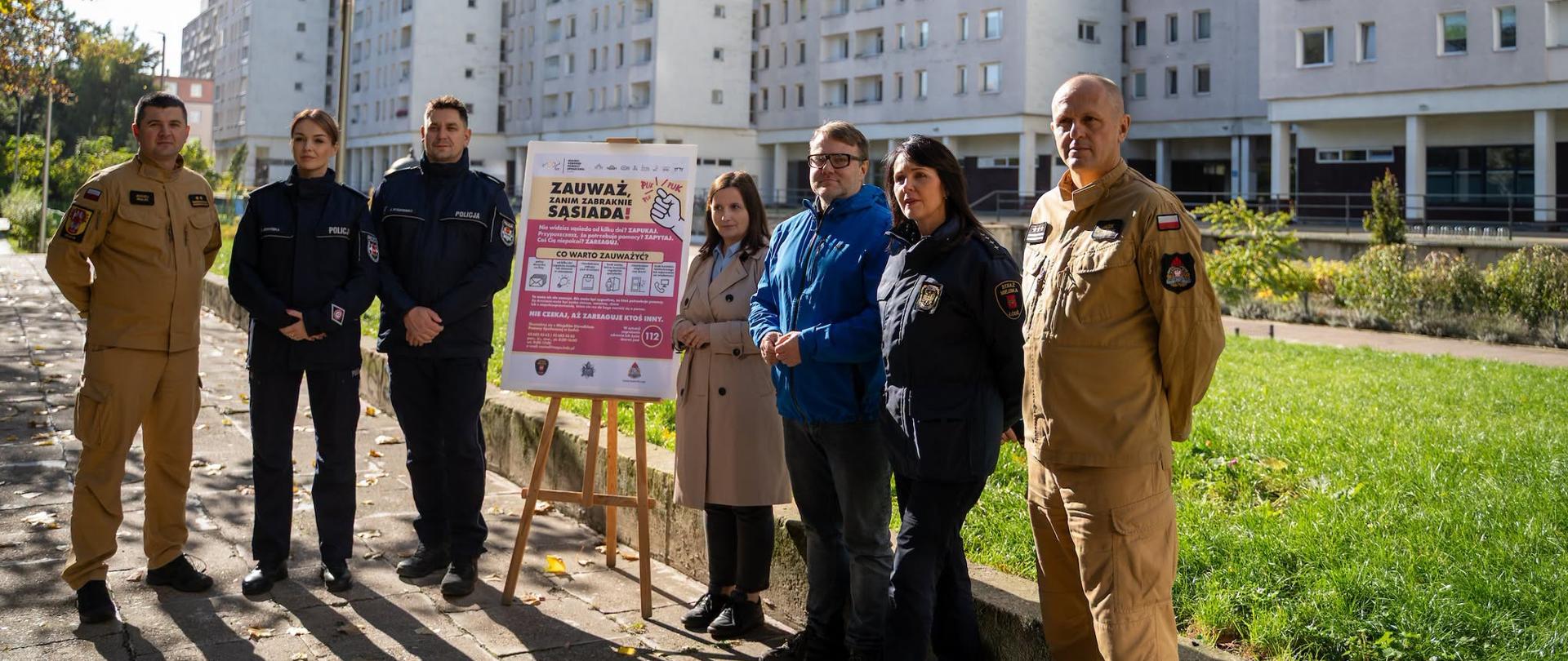 Przedstawiciele Państwowej Straży Pożarnej, Policji i Straży Miejskiej oraz organizatorzy kampanii „Zauważ, zanim zabraknie sąsiada” stoją przed budynkiem mieszkalnym obok plakatu promującego inicjatywę.