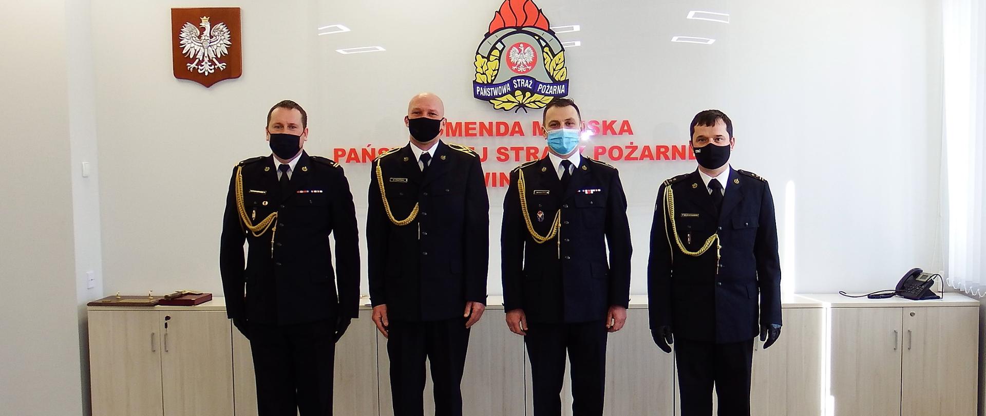 Wspólne zdjęcie emerytów z kierownictwem komendy. Czterech strażaków w mundurach wyjściowych ze sznurem en face. Od lewej stoją: mł. bryg. Tomasz Samborski, st. ogn. w stanie spoczynku Marcin Krużyński, asp. w stanie spoczynku Dariusz Kalemba, bryg. Tomasz Wojciechowski. W tle godło państwowe oraz godło PSP. 