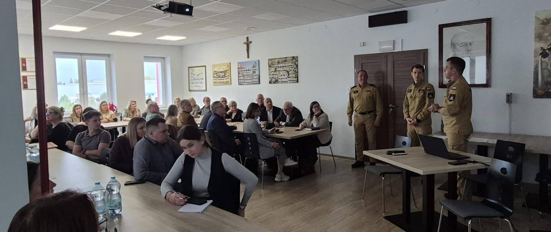 Na zdjęciu widać uczestników spotkania siedzących przy stołach oraz przedstawicieli Państwowej Straży Pożarnej stojących przed zgromadzonymi osobami. Uczestnicy słuchają wystąpienia, a strażacy przedstawiają omawiane zagadnienia.