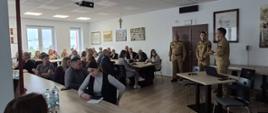 Na zdjęciu widać uczestników spotkania siedzących przy stołach oraz przedstawicieli Państwowej Straży Pożarnej stojących przed zgromadzonymi osobami. Uczestnicy słuchają wystąpienia, a strażacy przedstawiają omawiane zagadnienia.