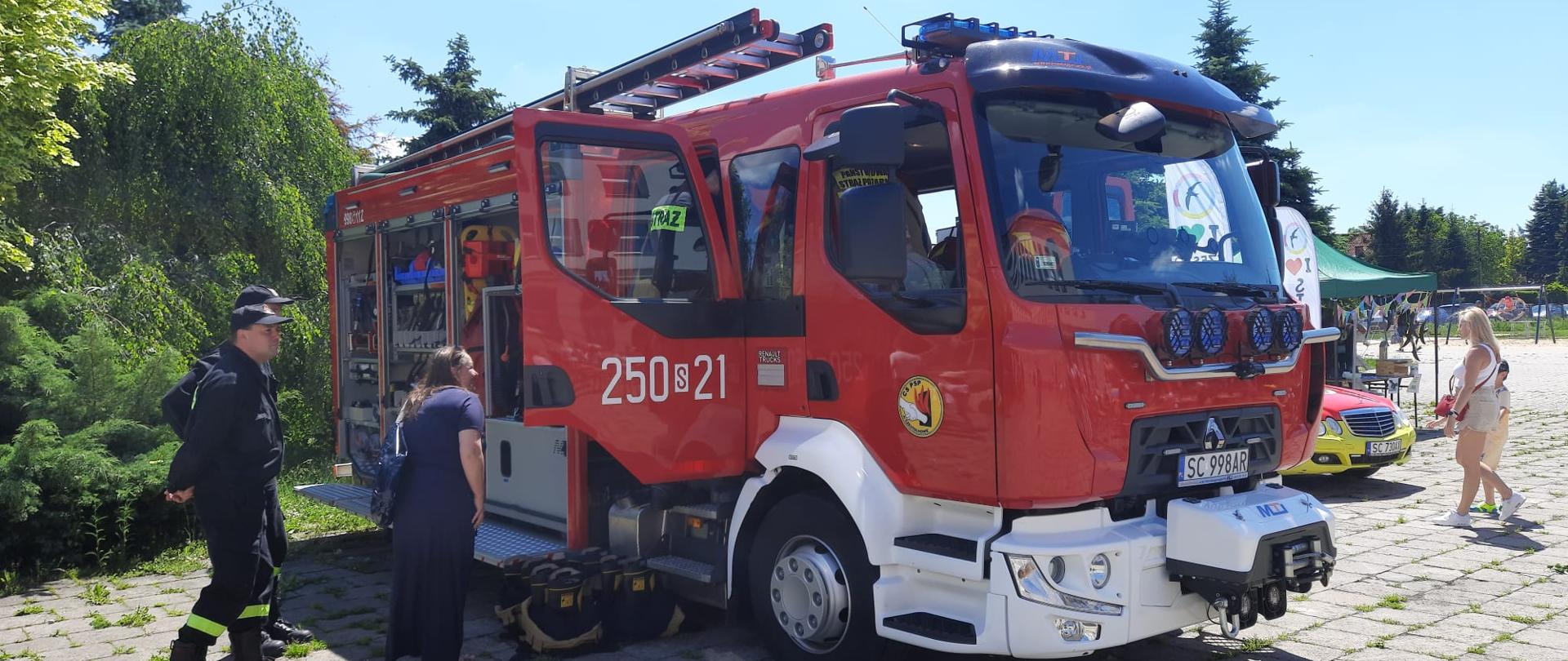 Na zdjęciu samochód gaśniczy stojący na parkingu podczas festynu, po lewej dwóch strażaków oraz kobieta