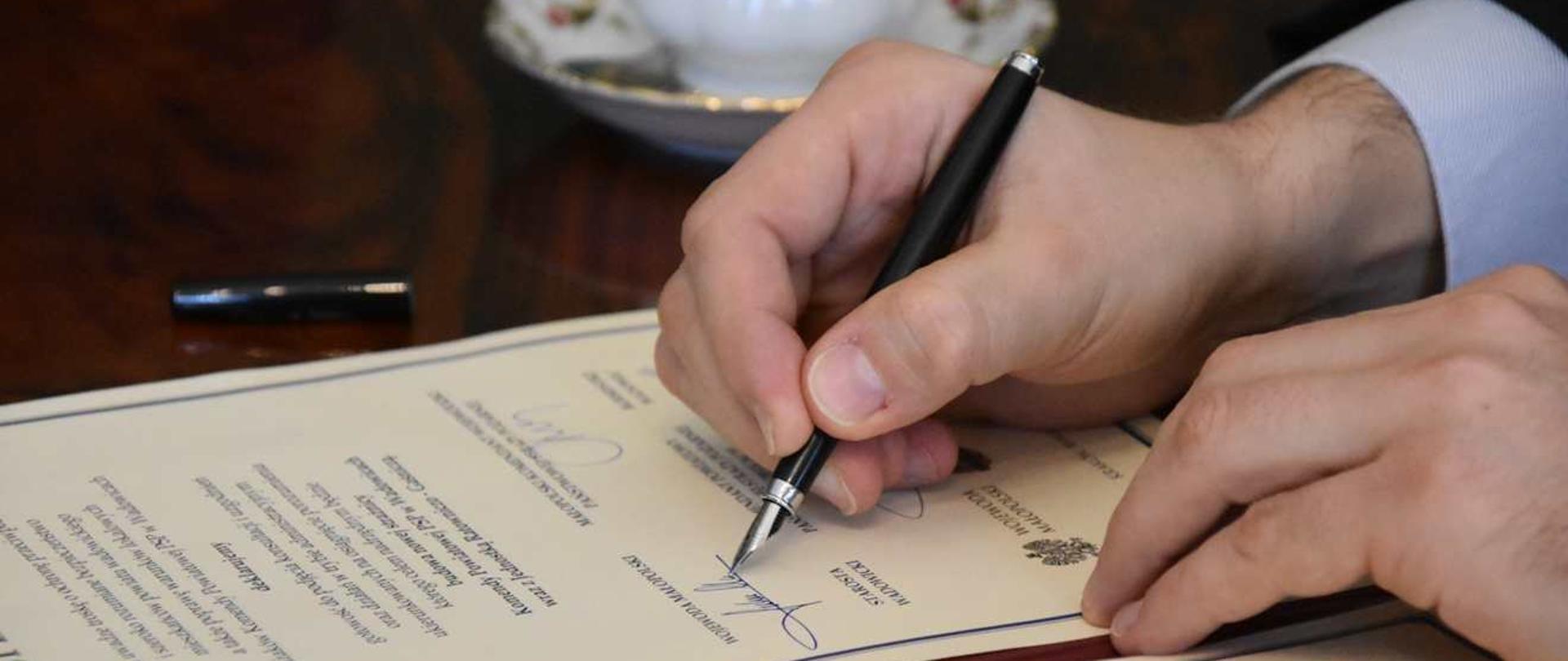 Widok na dokument. Po prawej stronie widoczne dłonie trzymające pióro. Widoczne wykonywanie jednego z podpisów na liście intencyjnym