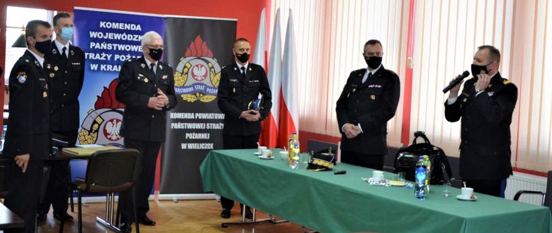 6 strażaków stoi ustawionych w półokręgu , pierwszy po prawej stronie - zastępca KG PSP przemawia, po prawej stronie stoi stół z zielonym obrusem