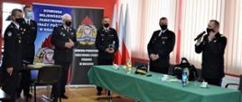 6 strażaków stoi ustawionych w półokręgu , pierwszy po prawej stronie - zastępca KG PSP przemawia, po prawej stronie stoi stół z zielonym obrusem