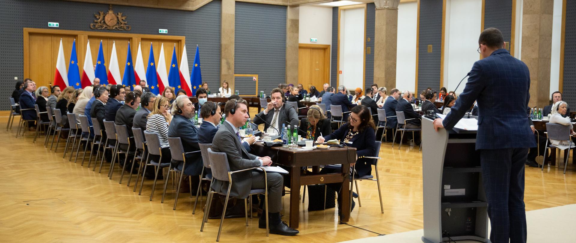 Duża sala z flagami Polski i Unii Europejskiej. Długie, ustawione równolegle dwa stoły wraz z ludźmi w słuchawkach na uszach.