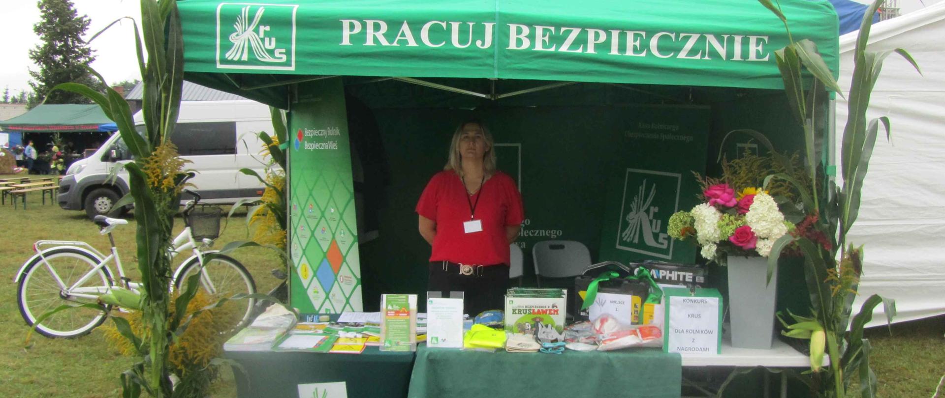 Stoisko informacyjne KRUS udekorowane kwiatami, Pod zielonym namiotem stoi kobieta – pracownik KRUS.