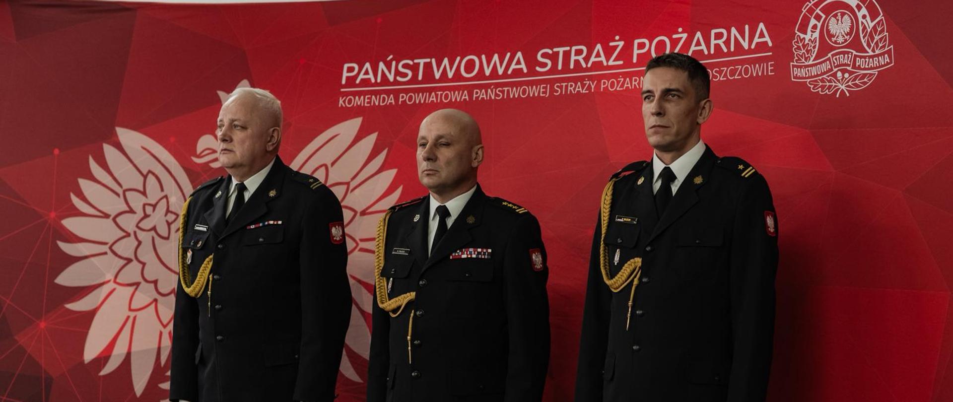 Trzech strażaków stoi na tle czerwonej ścianki strażackiej