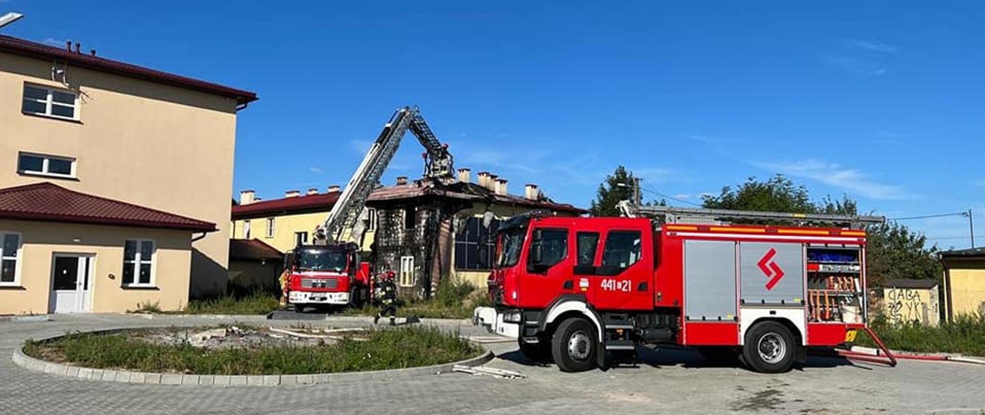 Na zdjęciu widać dwa czerwone samochody strażackie. Jeden z nich to podnośnik, który ustawiony jest przy palącym się budynku. Drugi z numerem 441E21 ma otwartą skrytkę i podłączone czerwone węże strażackie. W tle widać drugi budynek. Oba są jednopiętrowe. Jest widno.