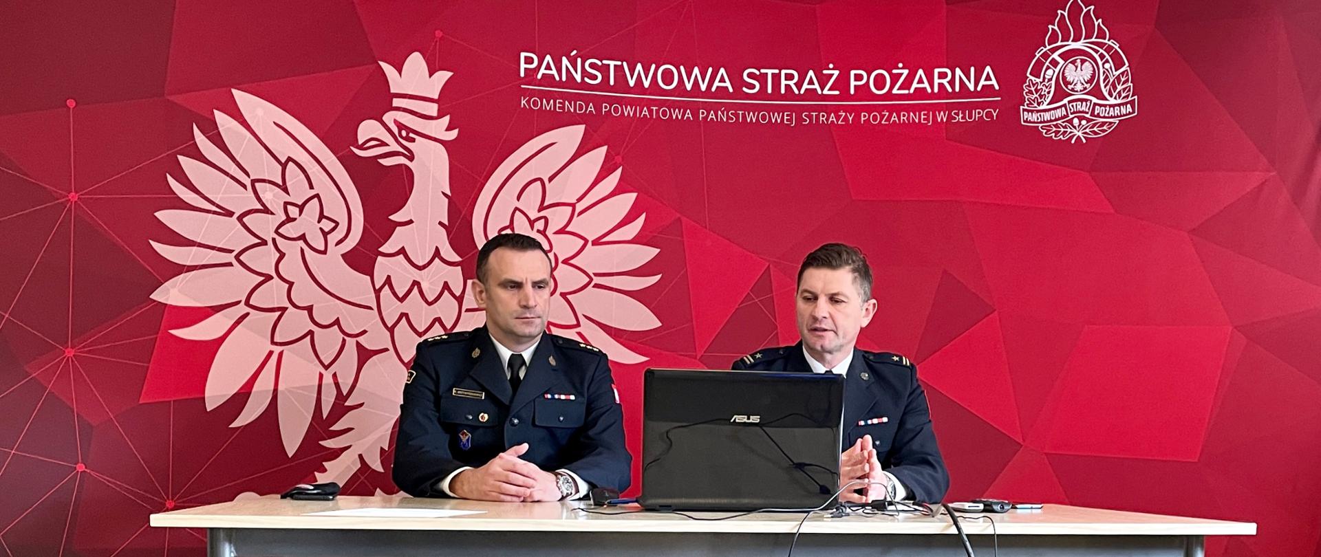 Komendant z zastępcą siedzą przy stole na który znajduje się otwarty laptop za nimi baner z logiem PSP oraz godłem na bordowym tle 