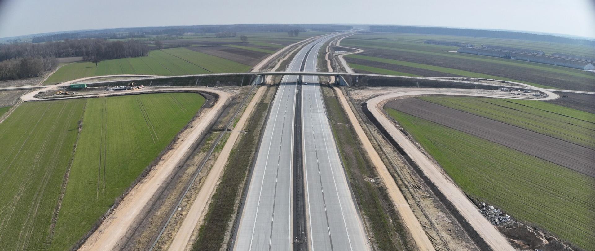 Zdjęcie przedstawia widok z lotu ptaka na szeroki, nowoczesny plac budowy autostrady A2, przecinający płaski, rolniczy krajobraz. W centrum kadru biegną dwie główne, równoległe jezdnie o jasnoszarej nawierzchni, oddzielone pasem rozdziału. W oddali widać gotowy wiadukt drogowy, który przebiega prostopadle nad autostradą.
Po obu stronach głównych jezdni znajdują się piaszczyste drogi serwisowe oraz szerokie pasy wyrównanej ziemi, przygotowane pod dalsze prace. Po lewej i prawej stronie drogi rozciągają się zielone i brązowe pola uprawne, gdzieniegdzie poprzecinane kępami drzew. Perspektywa zbieżna sprawia, że trasa zdaje się ciągnąć aż po horyzont pod jasnym, lekko zamglonym niebem.