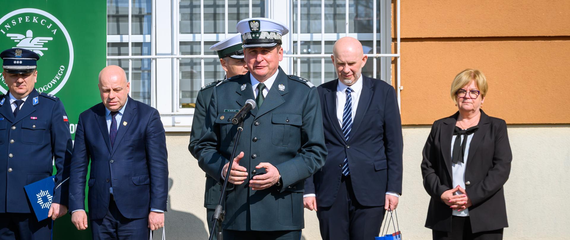 Mężczyzna w mundurze przemawia do mikrofonu. Za nim stoją ludzie. 