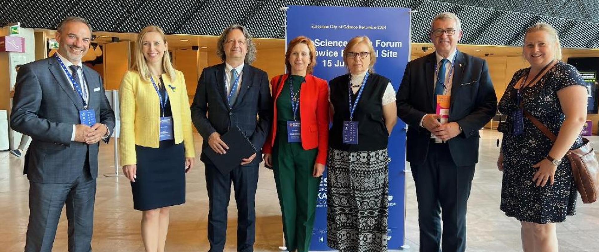 Grupa osób stoi na środku dużej sali, za nimi plakat z napisem EuroScience Open Forum.