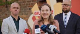 Minister funduszy i polityki regionalnej Katarzyna Pełczyńska-Nałęcz stoi przed mikrofonami dziennikarzy i się uśmiecha
