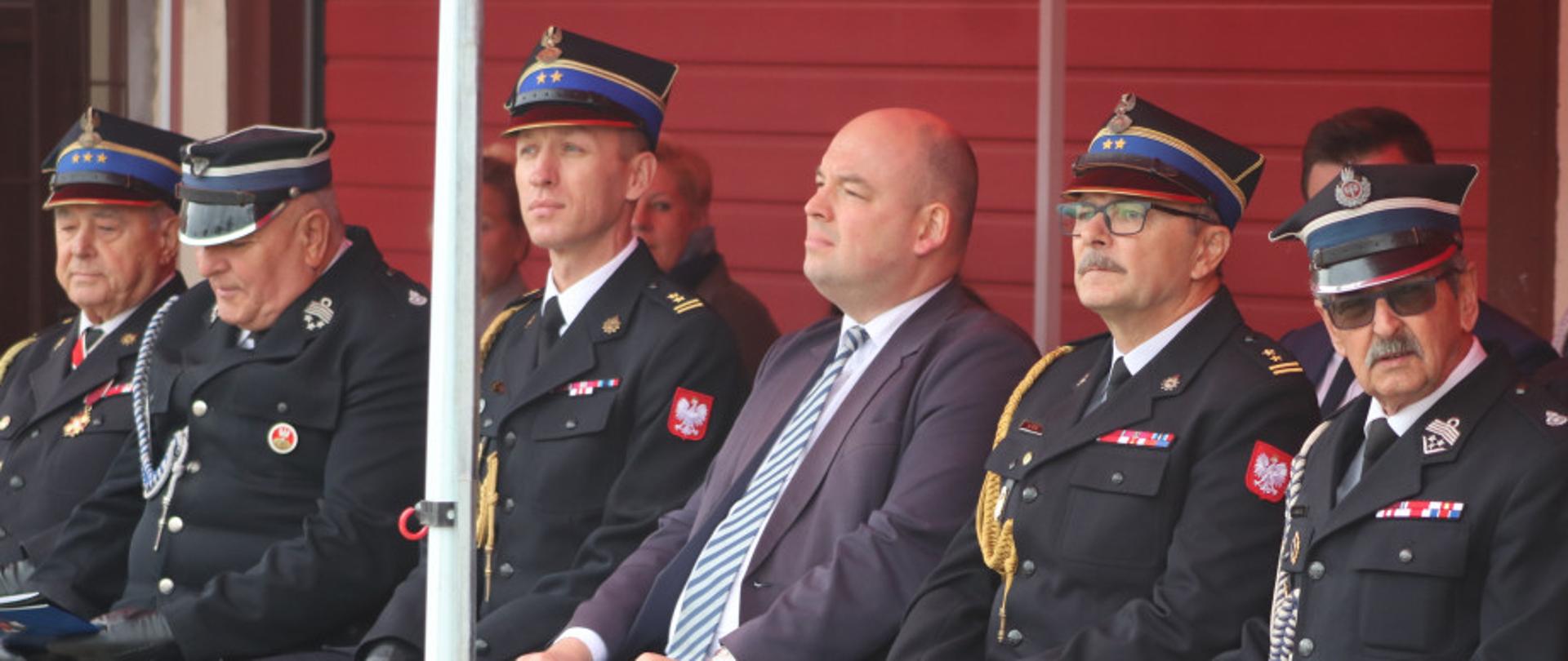strażacy w mundurach i w rogatywkach siedzą pod namiotem 