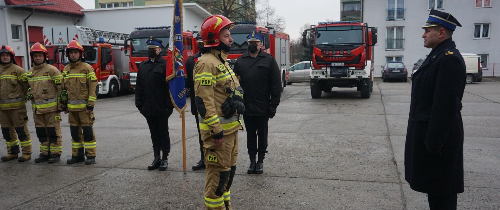Dzień. Plac wewnętrzny KP PSP w Zgorzelcu. Z lewej strony widać ustawionych w szeregu 3 strażaków ubranych w ubrania specjalne w kolorze piaskowym oraz czerwone hełmy. Obok nich stoi poczet sztandarowy KP PSP w Zgorzelcu. Funkcjonariusze w w poczcie ubranie w kurtko służbowe w kolorze czarnym oraz czapki rogatywki. Z prawej strony stoi Komendant Powiatowy PSP w Zgorzelcu ubrany w czarny płaszcz służbowy oraz czapkę rogatywkę. Przed Komendantem stoi strażak ubrany s ubranie specjalne w kolorze piaskowym oraz czerwony hełm. Strażak składa meldunek Komendantowi. Z strażakami w tle widać ustawione w szeregu 4 samochody ratowniczo-gaśnicze. 