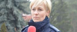 Przygotowywanie spotu edukacyjnego. Jedna z ulic w Dębnie Polskim. Na trawniku stoi policjantka. Trzymając w ręce mikrofon wypowiada swoją kwestię.